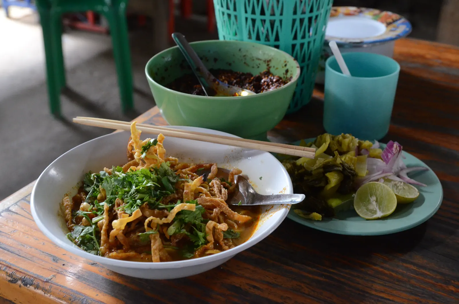 A bowl of khao soi with crispy noodles, pickled mustard greens, shallot and lime on a Chiang Mai shophouse table, chilli jar behind