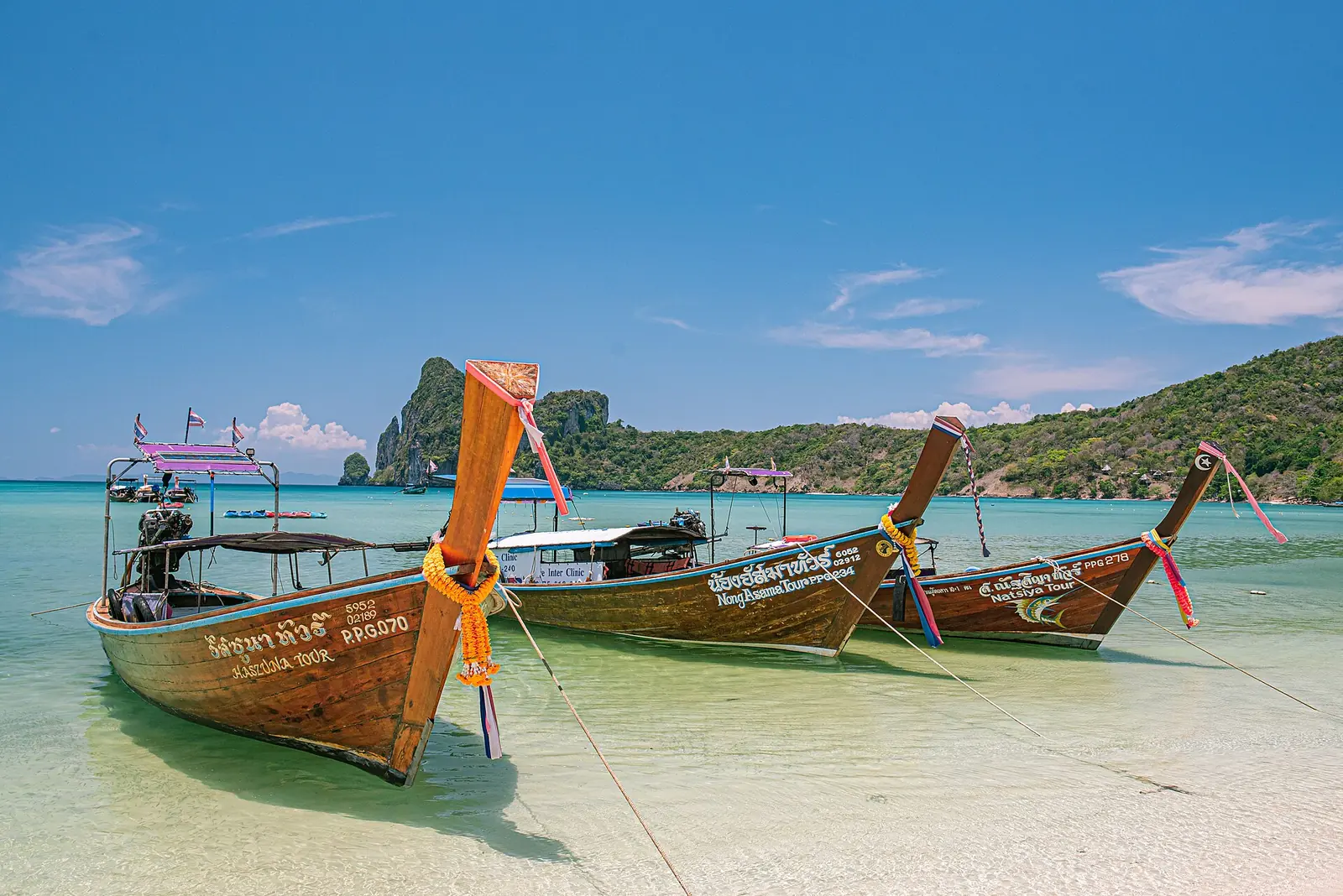Three wooden longtail boats with coloured ribbons moored in shallow turquoise water off a Krabi beach, limestone karst behind
