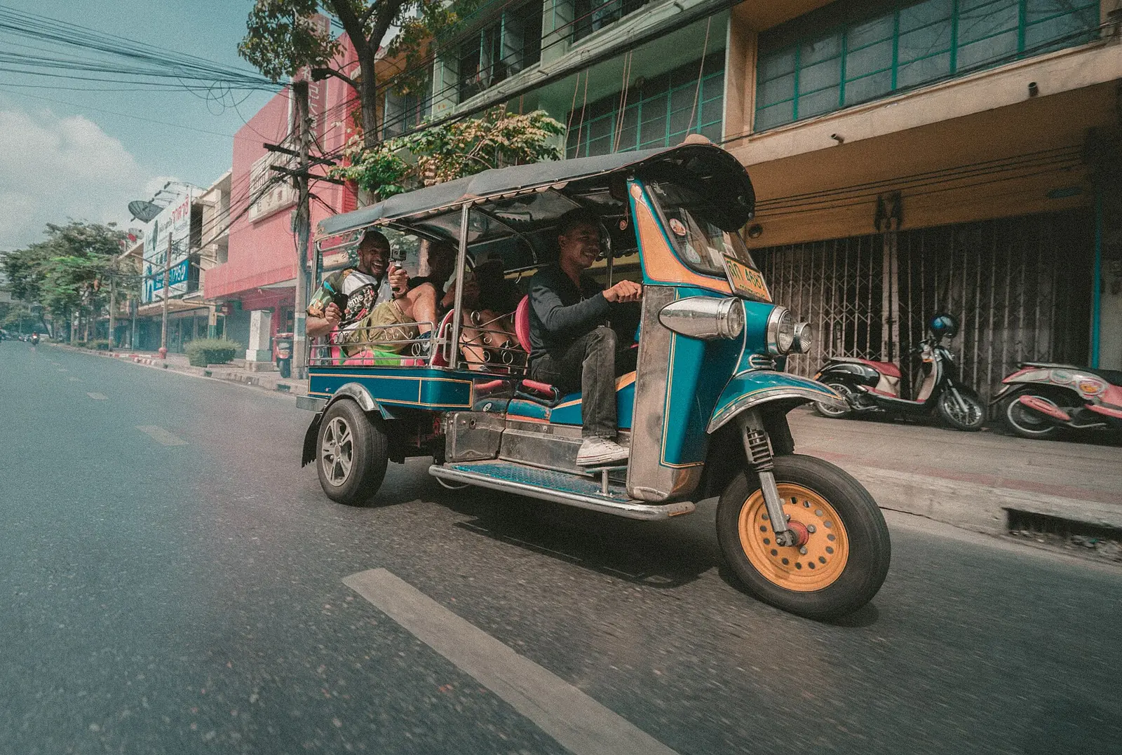 A blue tuk-tuk carrying passengers along a Bangkok shophouse street