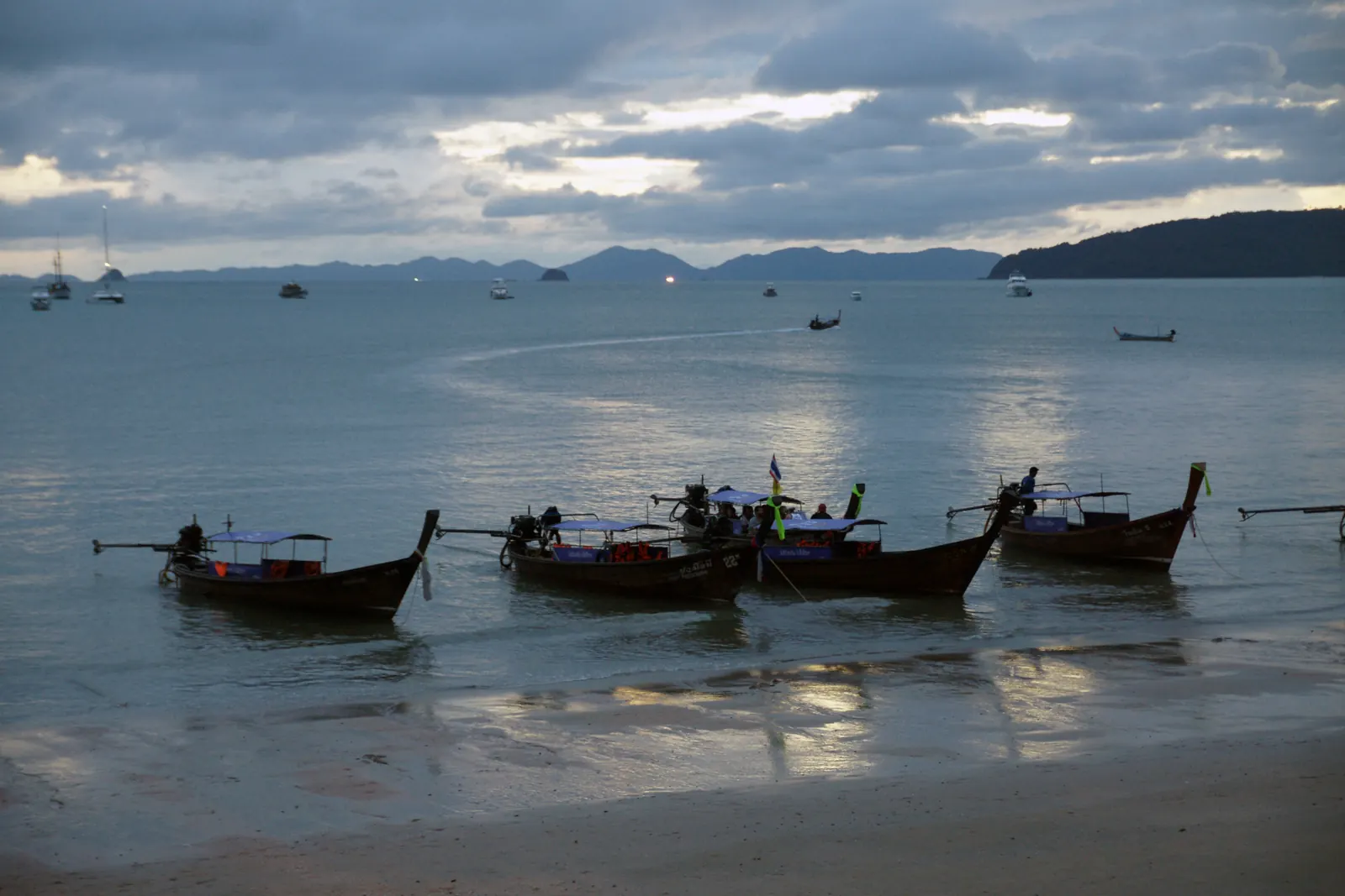 Longtail boats lined up on Ao Nang beach at dusk with the limestone headland behind, Krabi