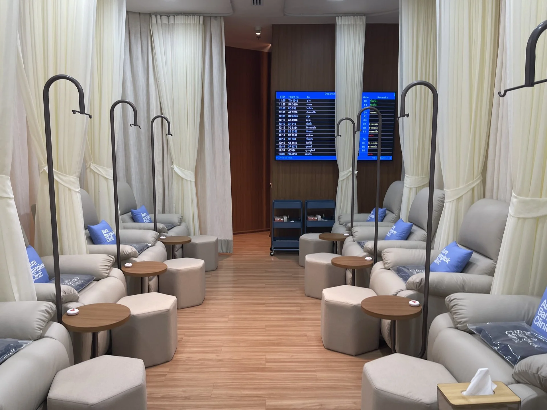 Row of IV drip recliners with a departures board at Aura Bangkok Clinic in Suvarnabhumi Airport Concourse G