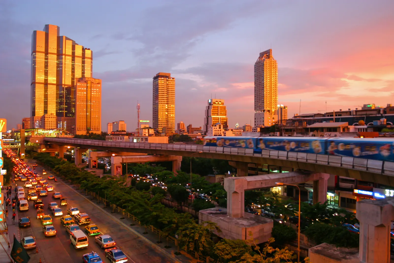 A BTS Skytrain on its elevated viaduct above a Bangkok road at sunset, with towers behind